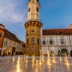 Beleuchteter Hauptplatz mit dem historischen Rathausturm von Bad Radkersburg und den leuchtenden Wasserspielen am Abend – stimmungsvolle Altstadt im Thermen- & Vulkanland Steiermark.