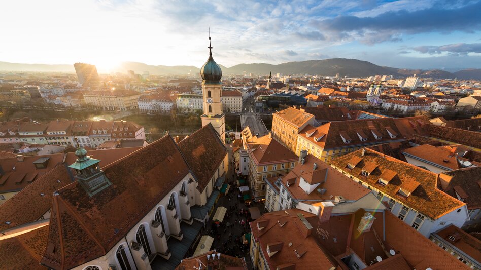Blick über die roten Dächer der Grazer Altstadt mit der Franziskanerkirche im Abendlicht.