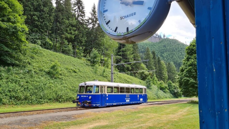 Im Vordergrund eine Uhr und dahinter fährt die Erzbergbahn