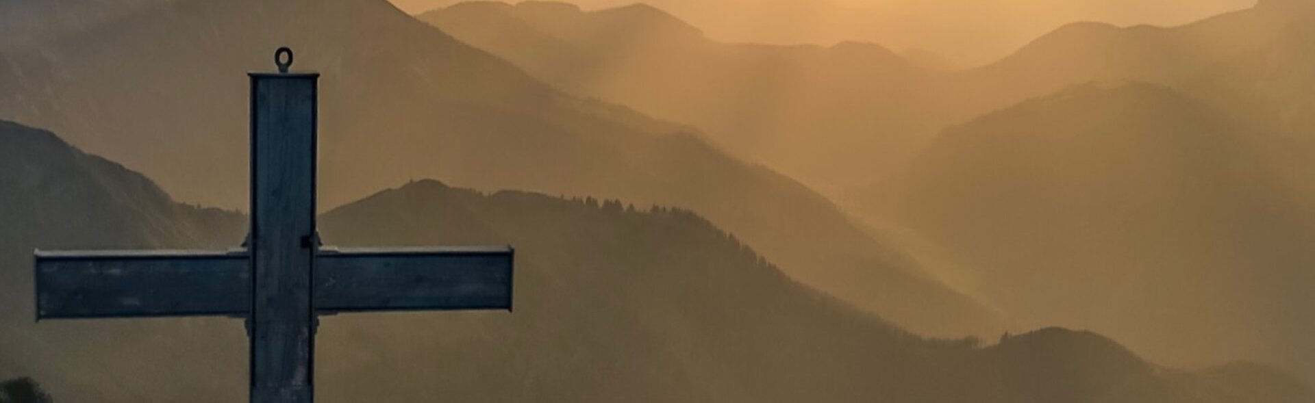 Eine Frau, die neben einem Gipfelkreuz steht und in den Sonnenuntergang hinter den Wolken blickt. 
