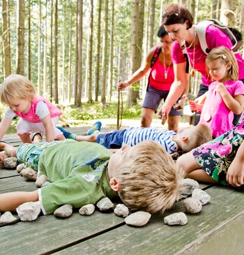 Zwei Kinder liegen auf einer Holzliege im Wald, Kinder legen Steine rundherum