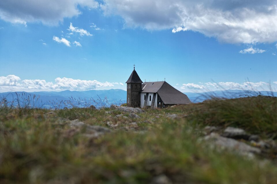 Eine kleine Kirche steht auf einem grünen Hügel. Im Hintergrund sind Wolken und Berge sichtbar. | © Isabella Painhapp