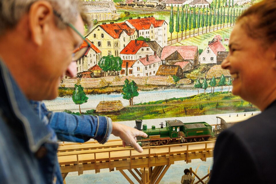 Zwei Personen betrachten eine Modelleisenbahn vor einer bunten Hintergrundleinwand. Der Mann zeigt auf die Lokomotive, während die Frau lächelt. | © Wolfgang Speckner