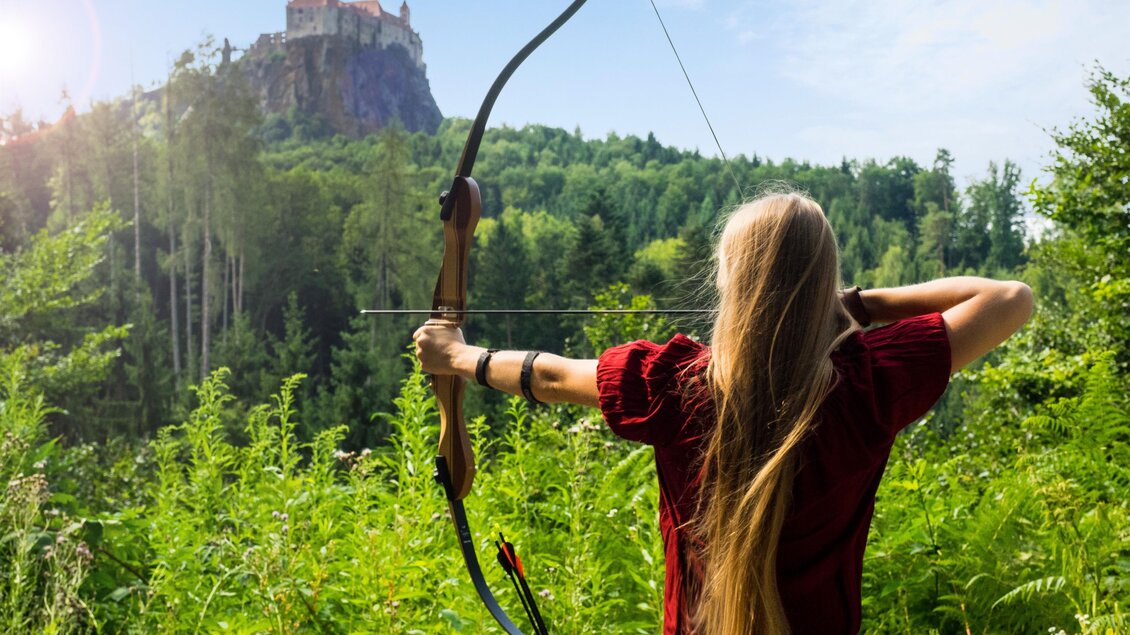 Eine Person mit langen Haaren zielt mit einem Bogen auf ein entferntes Schloss. Die Umgebung ist grün und sonnig, umgeben von Bäumen und Pflanzen. | © Naturbursch Training Riegersburg