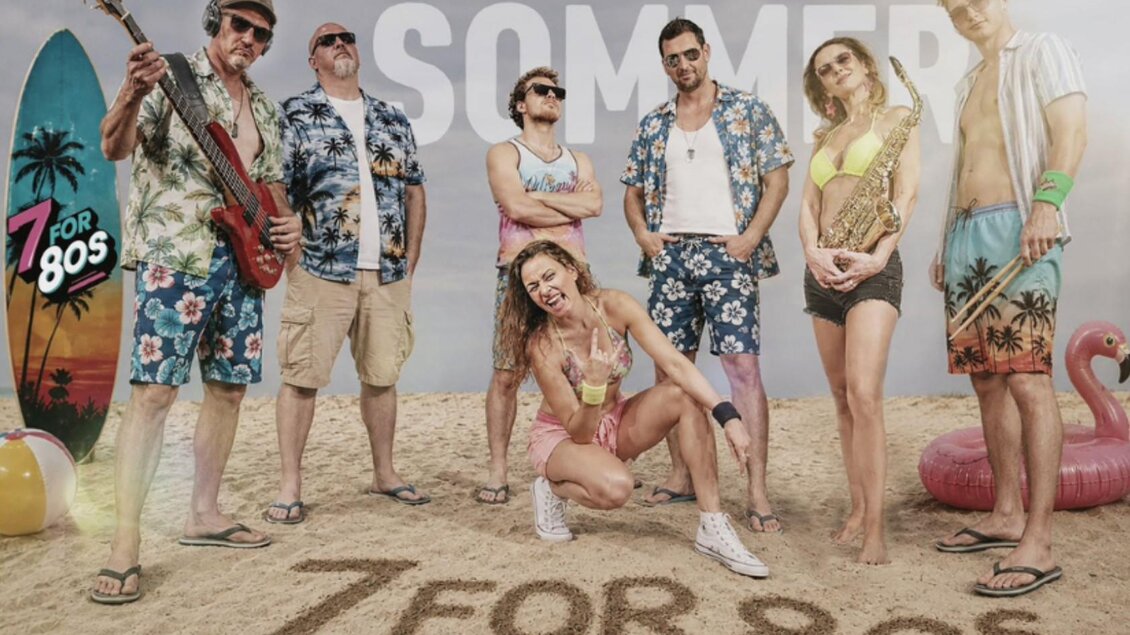 Eine Gruppe von Menschen in bunten Sommeroutfits steht am Strand. Im Hintergrund sind Strandutensilien und der Schriftzug "SOMMER" zu sehen. | © Inez Grieshofer