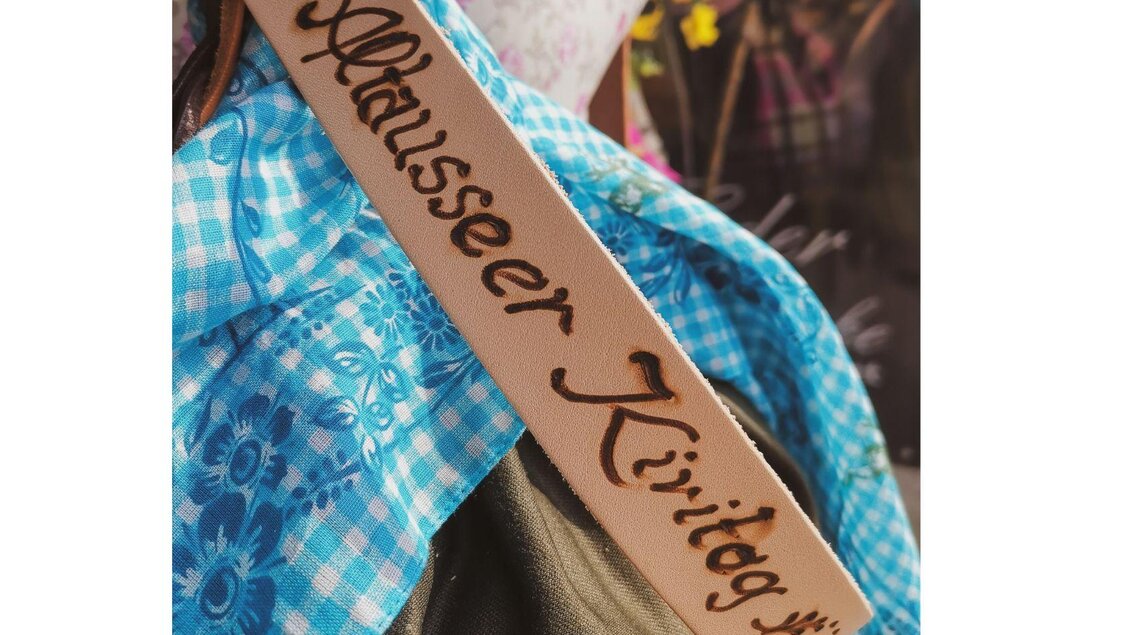 <p>Vor einem blau karierten Tuch ist eine Lederschärpe. Auf der Schärpe wurde "Altausseer Kiritog" in kunstvoller Schrift eingebrannt.</p> | © TVB Ausseerland Salzkammergut_Theresa Schwaiger