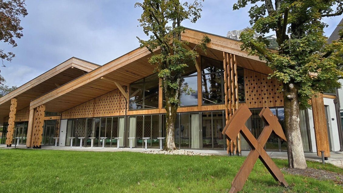 Der moderne Veranstaltungssaal im Herzen von Altaussee aus viel Holz und Glas. Im Vordergrund ist der grüne Kurpark der Gemeinde am Altausseer See zu sehen, der von einer Skulptur mit dem Bergmannszeichen aus Cortenstahl in seiner charakteristischen rostbraunen Farbe geschmückt wird, welche am rechten Bildrand zu sehen ist. | © Stephanie Bor