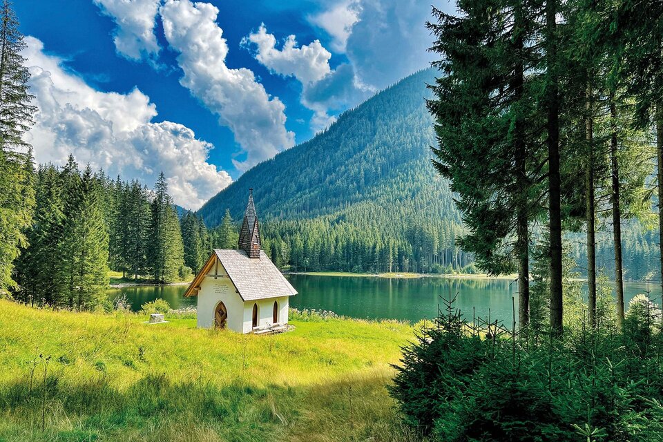 Eine malerische Landschaft mit einem kleinen Haus amSee. Umgeben von hohen Bergen und dichten Wäldern unter einem blauen Himmel mit Wolken. | © (c)KLOSTERWALD