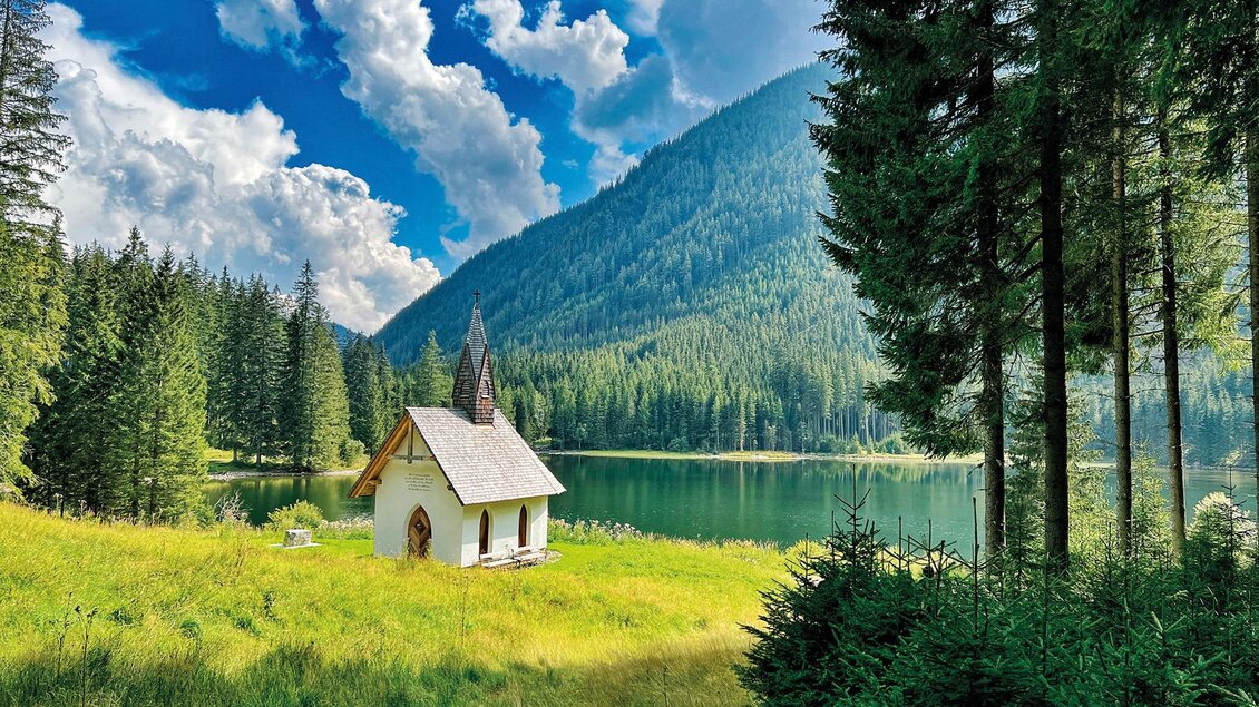 Eine malerische Landschaft mit einem kleinen Haus amSee. Umgeben von hohen Bergen und dichten Wäldern unter einem blauen Himmel mit Wolken. | © (c)KLOSTERWALD