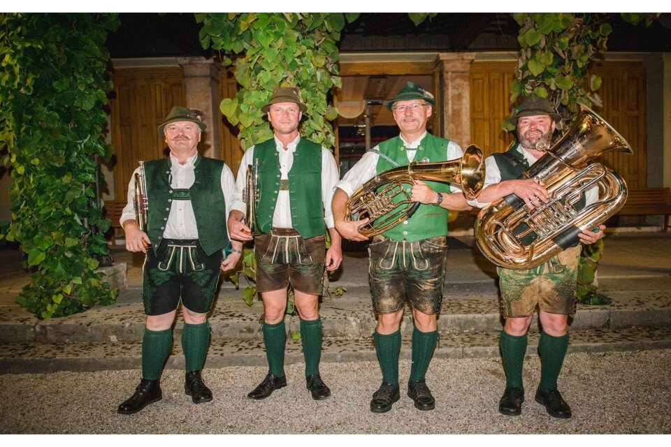 Eine Gruppe von vier Musikern in traditionellen Trachten steht vor einem historischen Hintergrund. Sie halten verschiedene Musikinstrumente, darunter eine Tuba. | © G.Baumann
