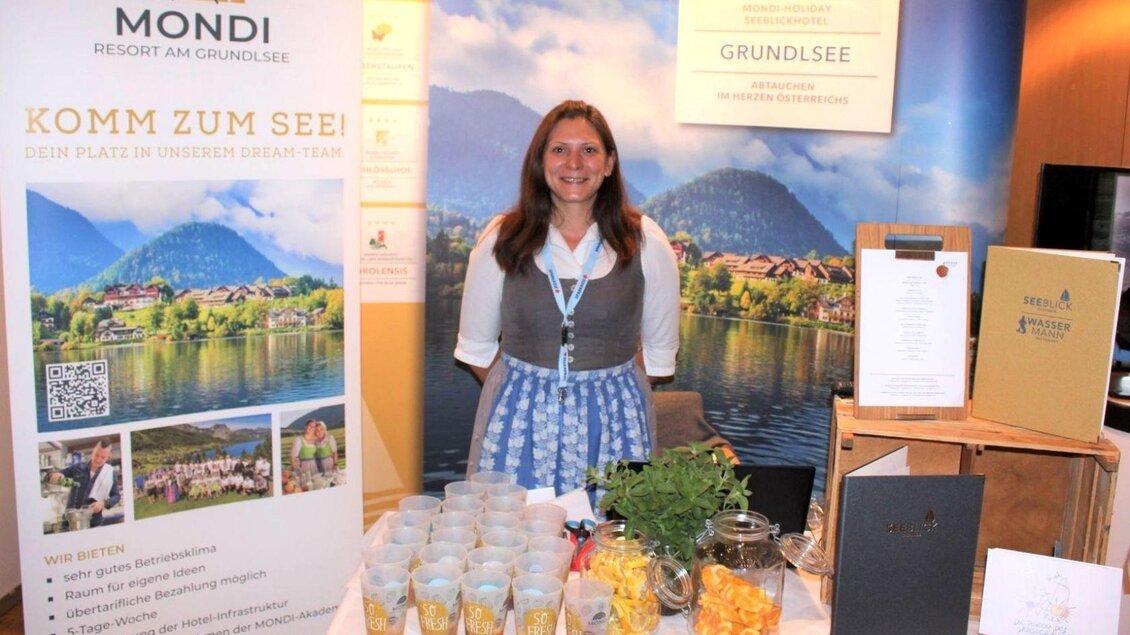 <p>Eine Frau steht beim Stand und informiert über das Mondi Resort am Grundlsee. Auf dem Tisch befinden sich Getränke und Snacks.</p> | © Arbeitsmarktservice Liezen, www.ams.at