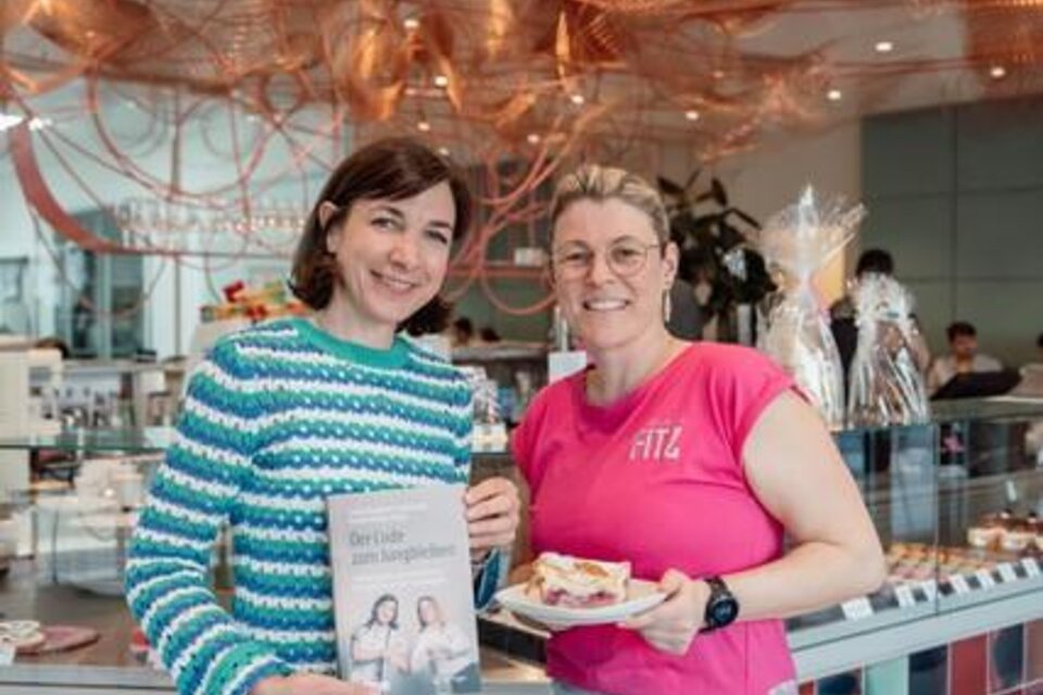 Zwei Frauen stehen in einem modernen Café und halten ein Kochbuch sowie eine Torte in den Händen. Im Hintergrund sind eine ansprechende Theke und dekorative Elemente zu sehen. | © SOSOCIAL