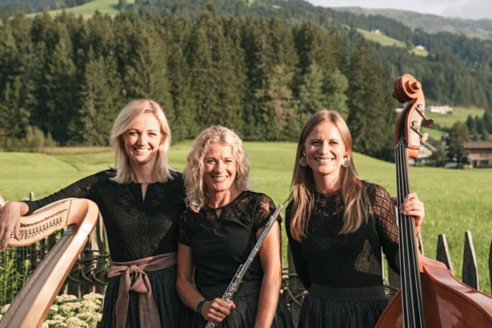 Drei Musikerinnen stehen in schwarz gekleideten Outfits in einer grünen Landschaft. Im Hintergrund sind Wälder und Berge zu sehen. | © Linortner