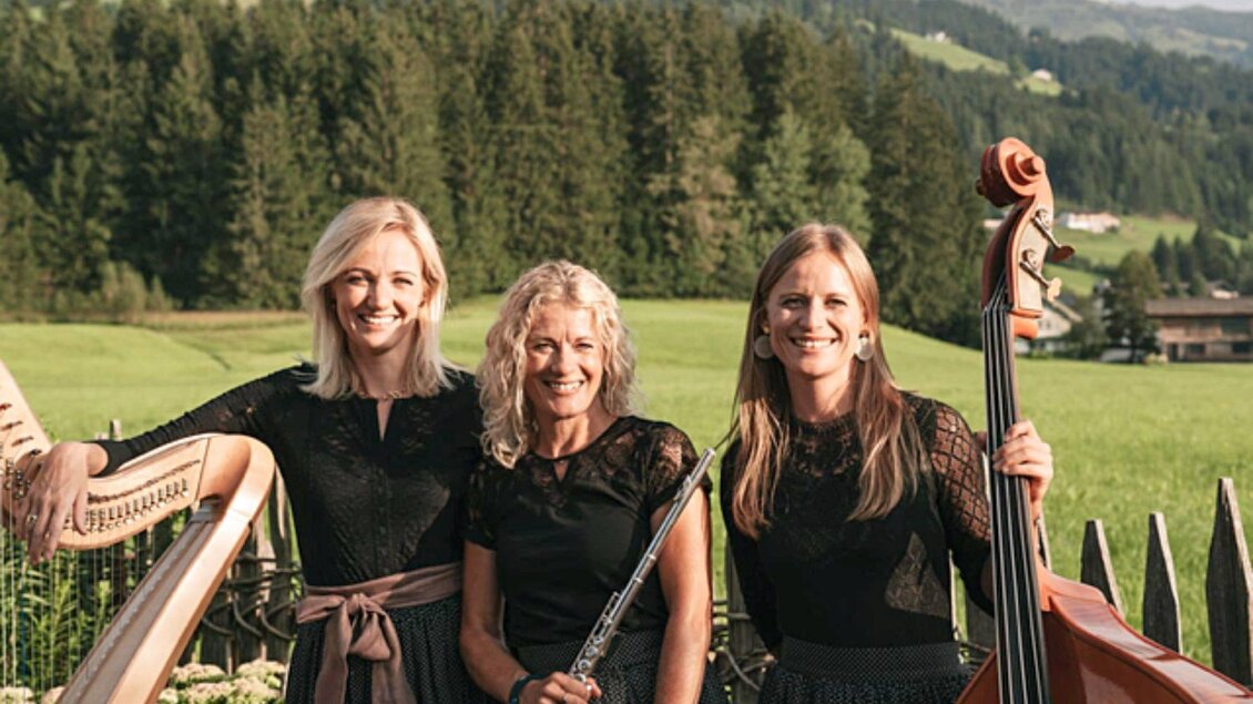 Drei Musikerinnen stehen in schwarz gekleideten Outfits in einer grünen Landschaft. Im Hintergrund sind Wälder und Berge zu sehen. | © Linortner
