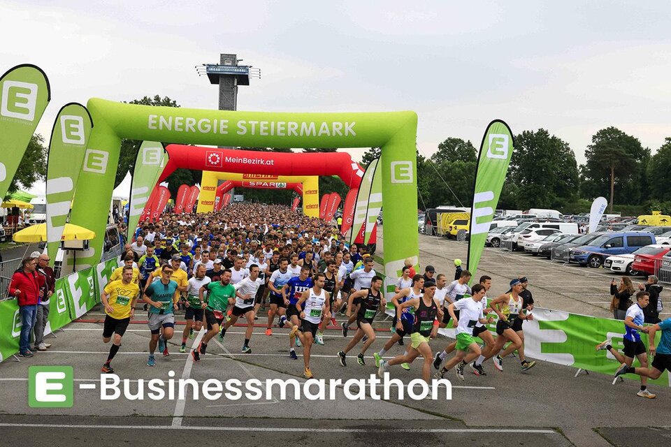 Ein großes Läuferfeld startet beim E-Businessmarathon unter einem bunten Zielbogen. Im Hintergrund sind viele Zuschauer und Fahrzeuge zu sehen. | © DieWoche