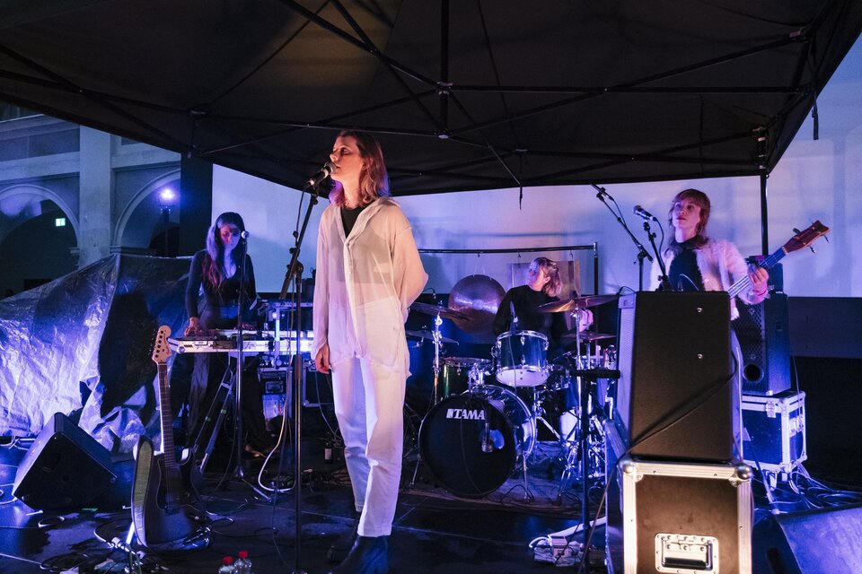 Eine Musikband tritt live auf einer Bühne auf. Die Sängerin in einem weißen Outfit steht im Mittelpunkt, während die anderen Bandmitglieder musizieren. | © -clara-wildberger