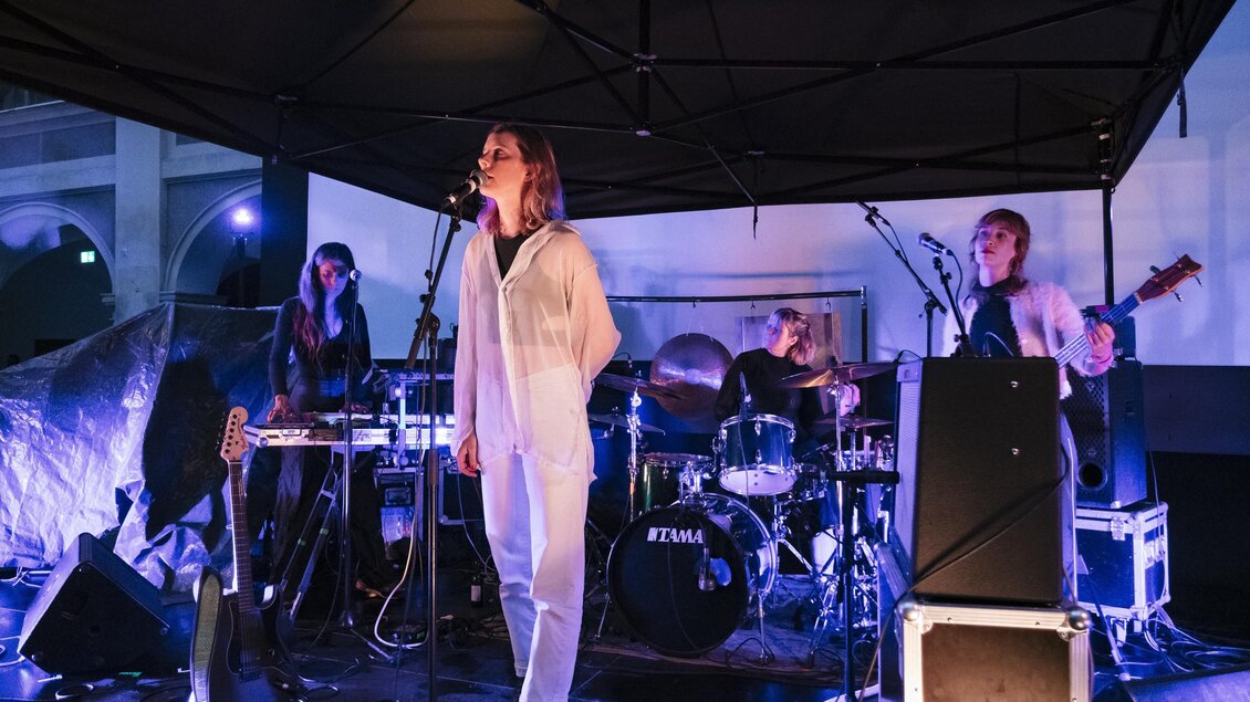 Eine Musikband tritt live auf einer Bühne auf. Die Sängerin in einem weißen Outfit steht im Mittelpunkt, während die anderen Bandmitglieder musizieren. | © -clara-wildberger