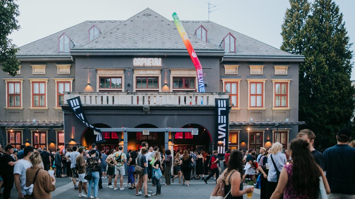 Ein lebhaftes Publikum versammelt sich vor einem historischen Gebäude. Bunte Fahnen wehen über dem Eingang, und die Atmosphäre ist festlich. | © -johanna-lamprecht