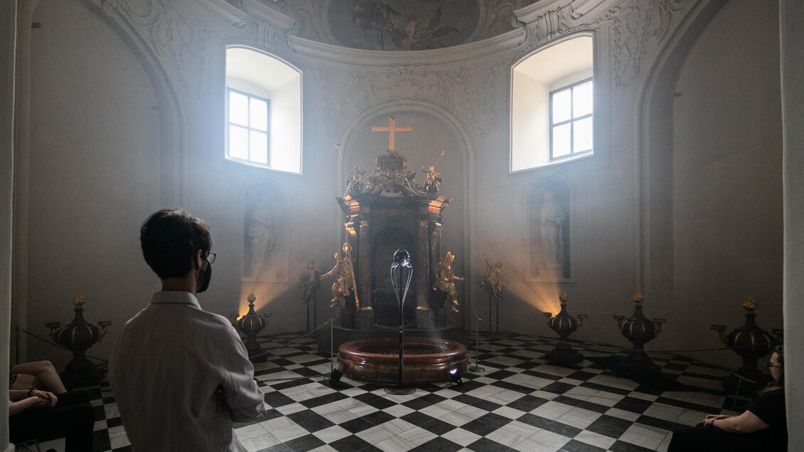 Ein innerer Raum mit einer Statue im Mittelpunkt, umgeben von schwachem Licht und Nebel. Zwei Personen stehen im Vordergrund und betrachten die Statue. | © johanna-lamprecht