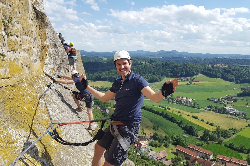 Ein Mann klettert an einer Felswand und lächelt in die Kamera. Im Hintergrund sind grüne Felder und ein blauer Himmel zu sehen. | © Naturbursch Training Riegersburg