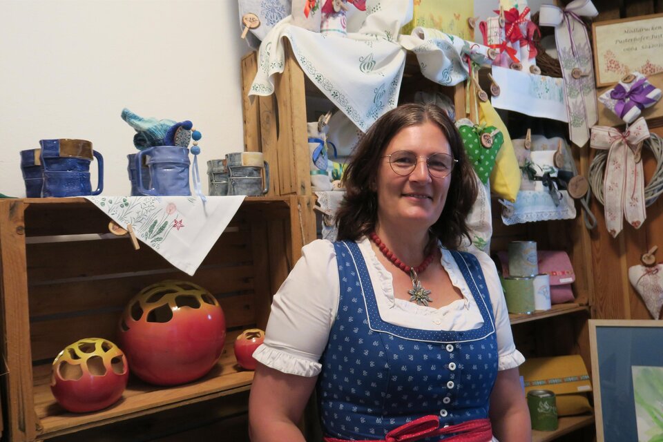 Eine Frau in traditioneller Tracht steht vor einem dekorierten Holzregal. Im Hintergrund sind handgefertigte Gegenstände und Dekorationen zu sehen. | © Marlene Zandl