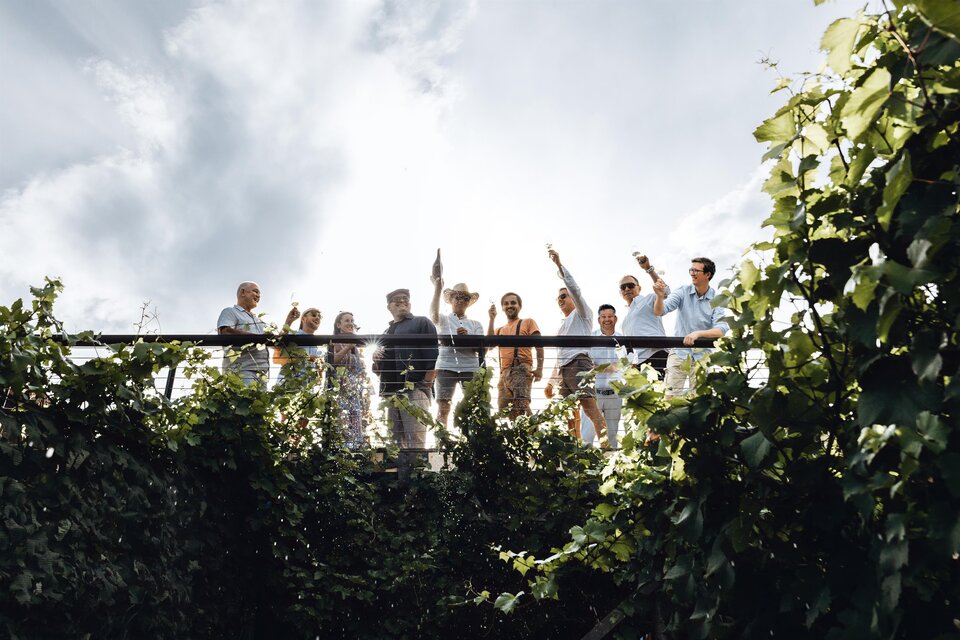 Eine Gruppe von Menschen steht auf einer Terrasse und winkt fröhlich. Dahinter sind grüne Pflanzen und ein bewölkter Himmel zu sehen. | © Katrin Meitz