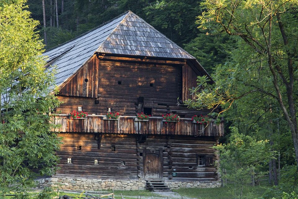 Ein rustikales Holzhaus umgeben von Bäumen. Der Balkon ist mit Blumen dekoriert und das Dach hat eine markante Form. | © Freilichtmuseum Stübing