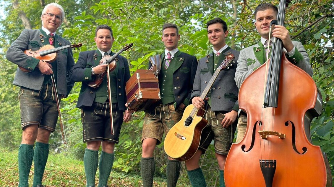 Eine Gruppe von fünf Musikern steht im Freien in traditioneller bayerischer Tracht. Sie halten verschiedene Instrumente wie Geige, Gitarre und Kontrabass in der Hand. | © Michael Weissensteiner