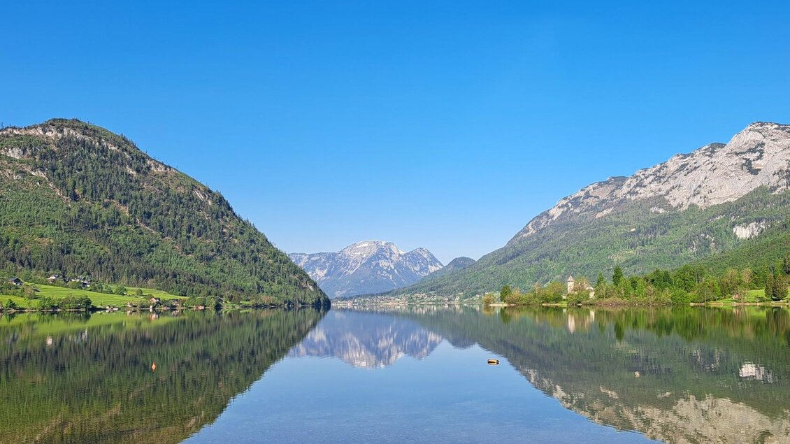 Ein ruhiger See, umgeben von hohen Bergen und grünem Wald. Der blaue Himmel spiegelt sich im Wasser. | © Franz Steinegger