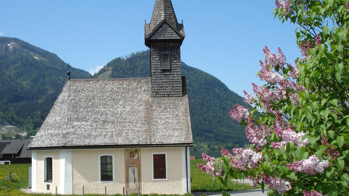 Eine malerische Kirche mit einem spitzen Turm, umgeben von Bergen. Im Vordergrund blühen lila Blumen. | © Franz Steinegger