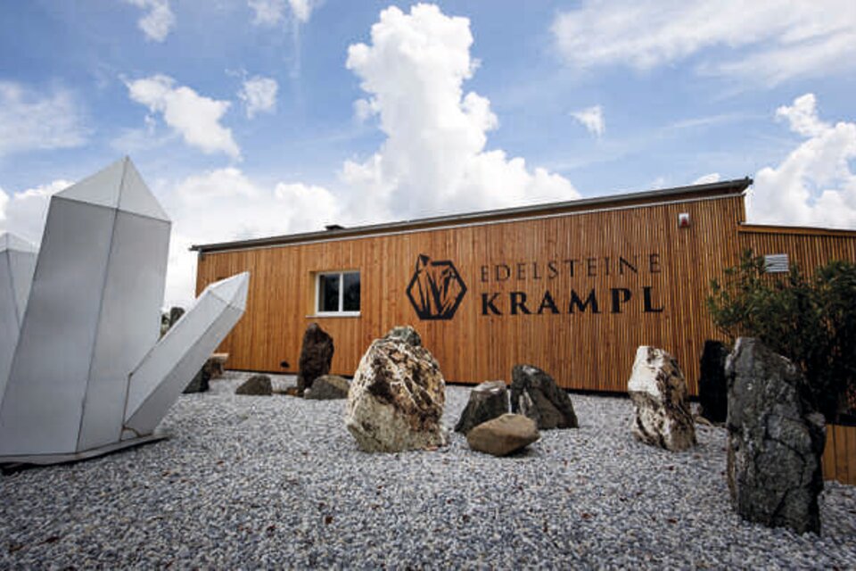 Ein modernes Holzgebäude mit dem Schriftzug "Edelsteine Kramp" ist vor einem klaren Himmel zu sehen. Im Vordergrund stehen große Steine und eine geometrische Skulptur. | © Edelsteine Krampl