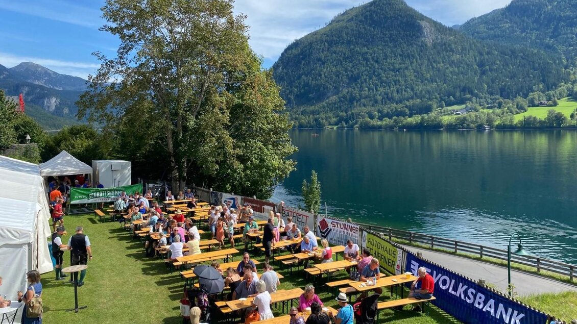 Eine belebte Uferpromenade mit langen Tischen und Sitzgelegenheiten am Wasser. Im Hintergrund sind Berge und ein klarer Himmel zu sehen. | © FF Grundlsee