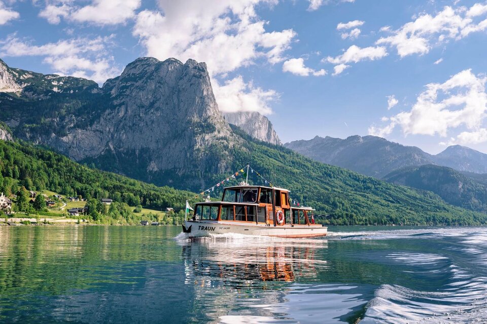 Ein Boot fährt ruhig über einen glitzernden See, umgeben von majestätischen Bergen. Der Himmel ist klar mit einigen weißen Wolken. | © Schifffahrt Grundlsee Toplitzsee