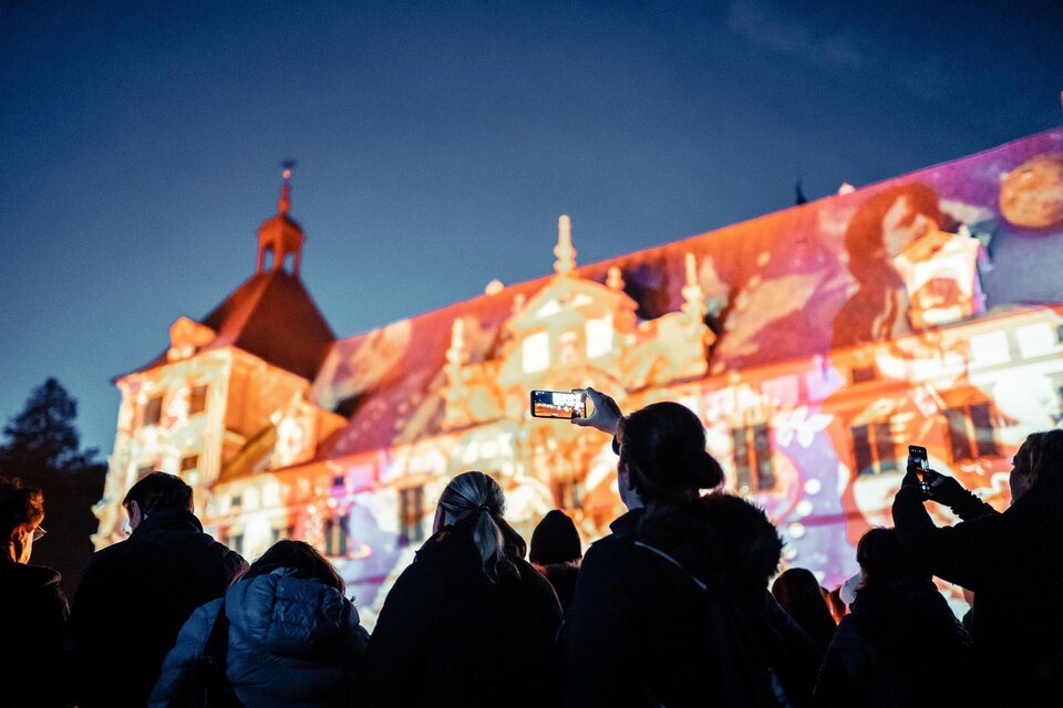 Eine Gruppe von Menschen steht vor einem Gebäude, das mit bunten Lichtern projiziert wird. Es ist Abend und die Stimmung ist festlich. | © (c) Alex Koch