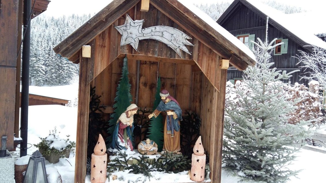 Eine Weihnachtskrippe aus Holz mit Figuren von Maria und Josef in einer verschneiten Landschaft. Umgeben von Tannenbäumen und Weihnachtsdekoration. | © Petra Kirchschlager
