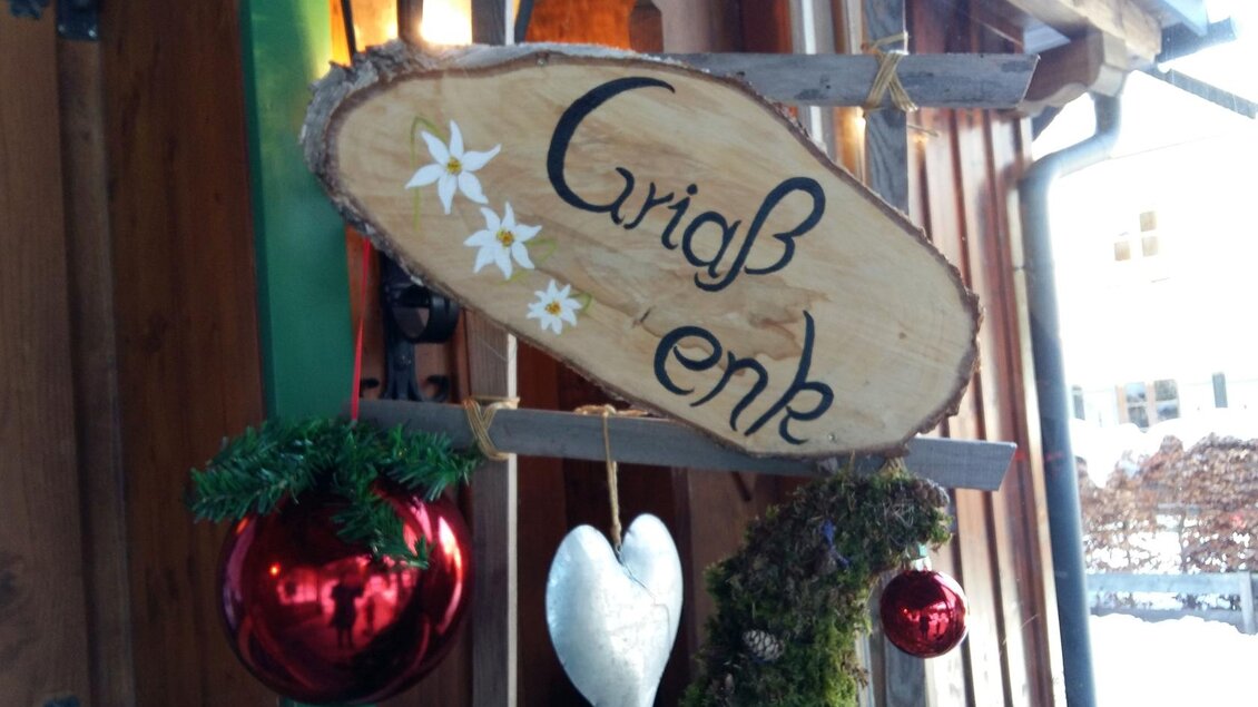 Ein handgemachtes Holzschild mit der Aufschrift "Griaß enk". Es ist mit einer Weihnachtsdekoration aus Tannenzweigen, roten Kugeln und einem weißen Herz geschmückt. | © Petra Kirchschlager