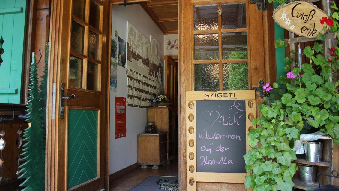 Ein einladender Eingang zu einem gemütlichen Gasthaus mit einer Tafel, die „Willkommen auf der Bloa-Alm“ zeigt. Der Bereich ist mit Pflanzen geschmückt und hat eine rustikale Atmosphäre. | © Viola Lechner