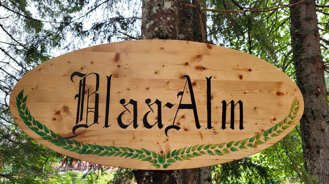 Ein Holzschild mit der Aufschrift „Blaa-Alm“. Es ist von grünen Blättern umrahmt und hängt zwischen Bäumen. | © Petra Kirchschlager