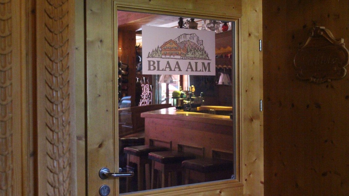 Eine Holzttür mit einem Fenster, das die Innenseite eines Gasthauses zeigt. Auf dem Fenster ist ein Schild mit der Aufschrift "BLAA ALM" zu sehen. | © Viola Lechner