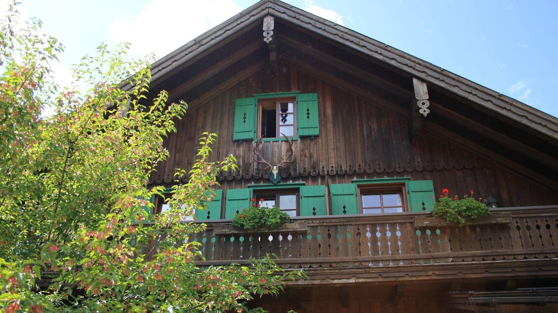 Ein traditionelles Holzhaus mit grünen Fensterläden und einem Balkon. Umgeben von Bäumen und blühenden Pflanzen. | © Viola Lechner