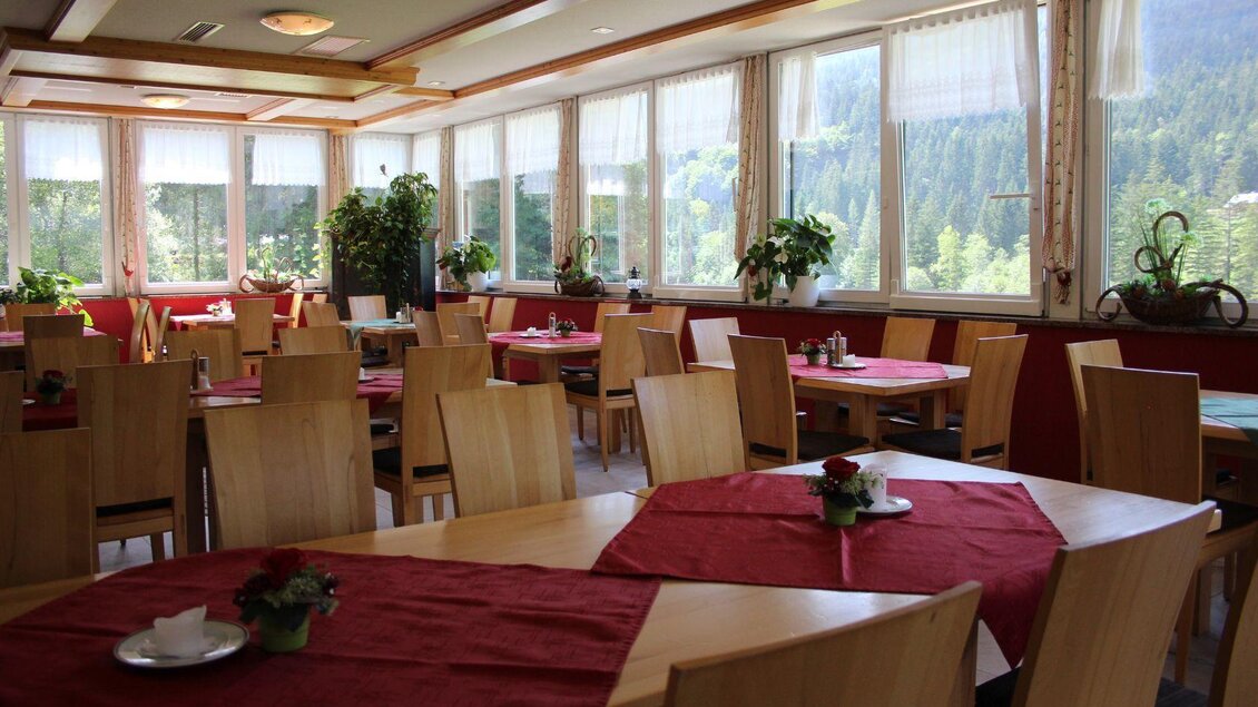 <p>Der einladende Wintergarten mit hellen Fenstern und Blick auf die Natur im Wirtshaus auf der Blaa Alm. Die Tische sind mit roten Tischdecken und kleinen Blumen dekoriert.</p> | © Viola Lechner