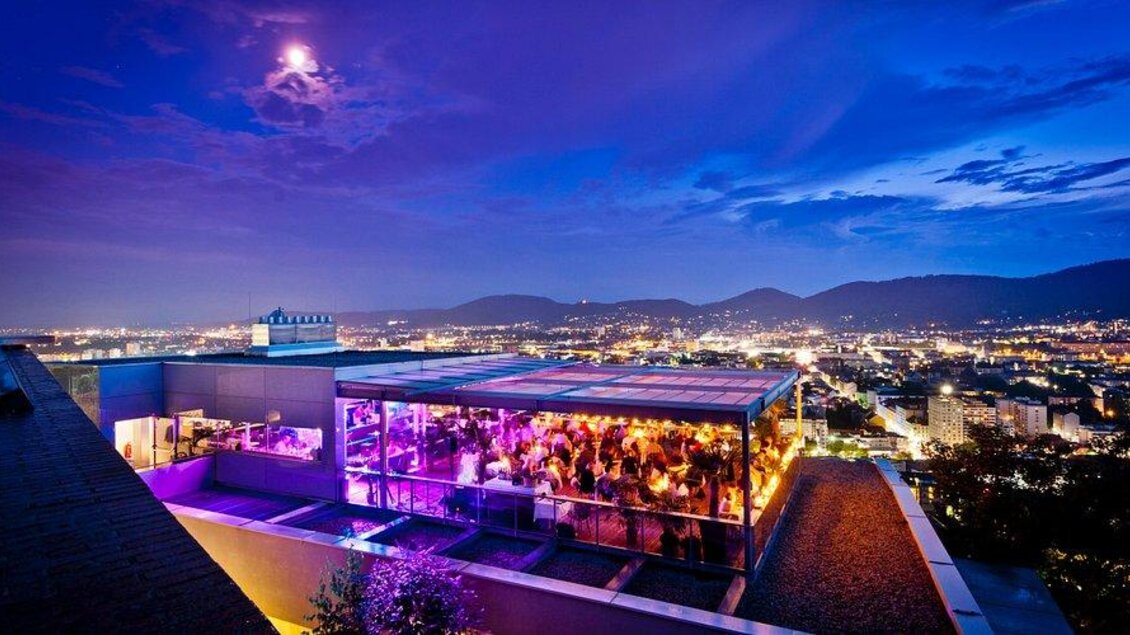 Eine stilvolle Rooftop-Bar mit bunten Lichtern und einer atemberaubenden Aussicht auf die Stadt bei Nacht. Der Himmel ist bewölkt und der Mond leuchtet über den Bergen. | © restaurant schlossberg