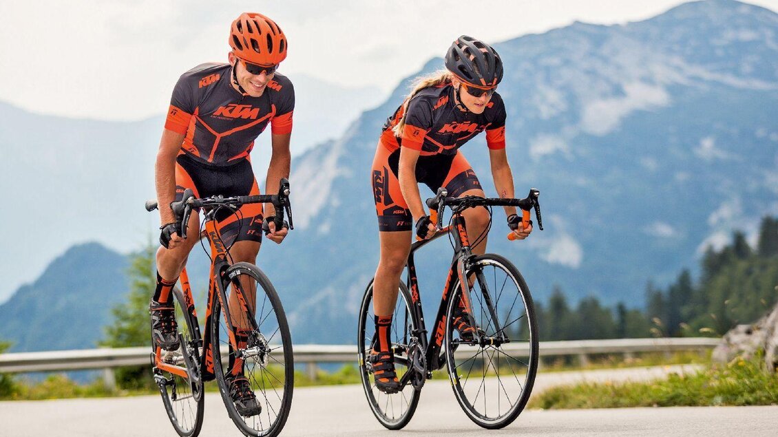 <p>Zwei Radfahrer in orange-schwarzen Trikots der Firma KTM fahren auf der Loser Panoramastraße. Im Hintergrund ist der Sandling zu sehen.</p> | © SkgtTrophy_HeikoMandl