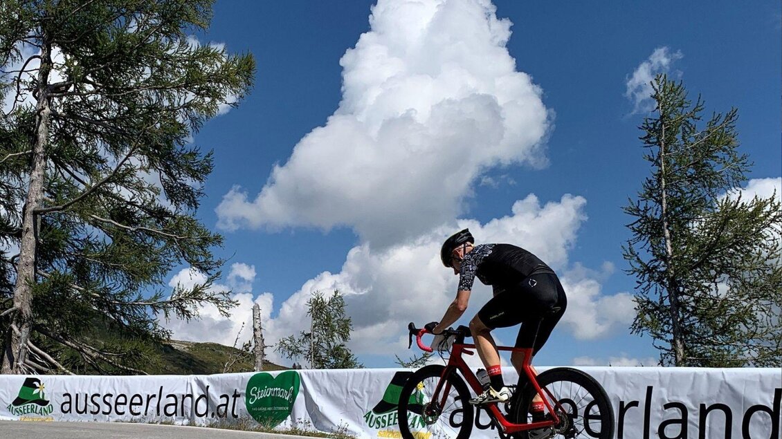 <p>Ein Radfahrer fährt entlang der Loser Panoramastraße unter einem klaren Himmel mit großen Wolken. Im Hintergrund sind Bäume und eine Werbung vom Tourismusverband Ausseerland zu sehen.</p> | © Lindpointner