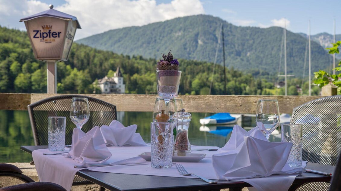 Ein elegant gedeckter Tisch im Dorfwirtshaus Stöckl mit Blick auf den wunderschönen Grundlsee und die umliegenden Berge des Ausseerlandes. Die Tischdekoration umfasst Gläser und Servietten, die schön angeordnet sind. | © Dorfwirtshaus Stöckl