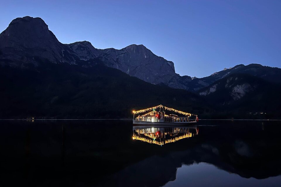 Ein ruhiger See mit einem beleuchteten Boot und majestätischen Bergen im Hintergrund. Der Himmel ist tiefblau und der Moment strahlt eine friedliche Stimmung aus. | © Schifffahrt Grundlsee Toplitzsee