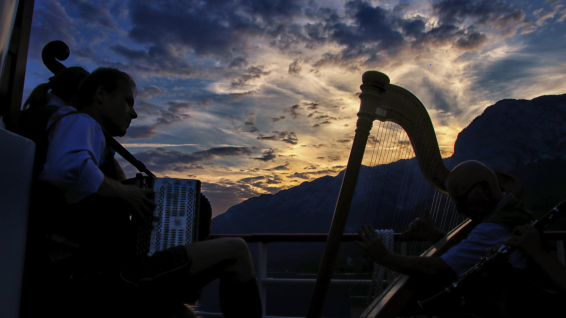 Eine Gruppe von Musikern spielt auf einem Boot bei Sonnenuntergang. Die Silhouetten sind vor einem dramatischen Himmel mit Bergen im Hintergrund zu sehen. | © Astrid Eder