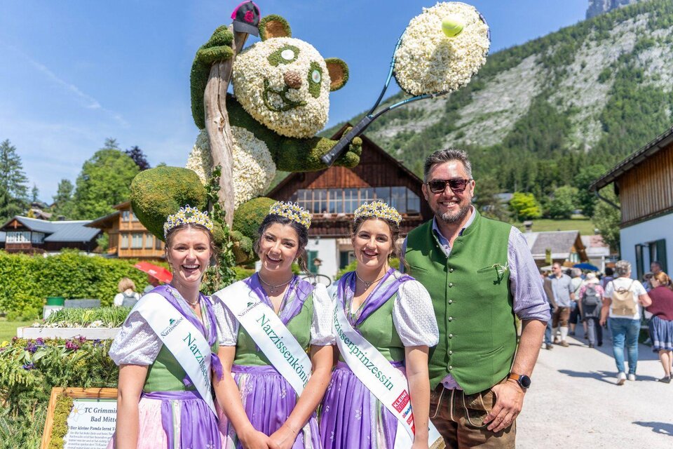 <p>Die drei gekrönten Narzissenhoheiten in traditionellen Trachten mit ihren schönen Kronen von den Narzissenwiesen stehen vor einer Narzissenfigur mit der schönen Landschaft im Hintergrund. | © Narzissenfestverein_Jakob Grill