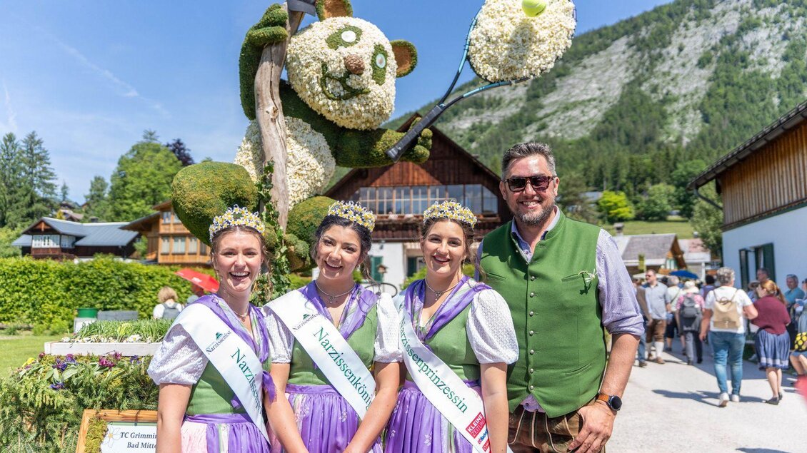 <p>Die drei gekrönten Narzissenhoheiten in traditionellen Trachten mit ihren schönen Kronen von den Narzissenwiesen stehen vor einer Narzissenfigur mit der schönen Landschaft im Hintergrund. | © Narzissenfestverein_Jakob Grill