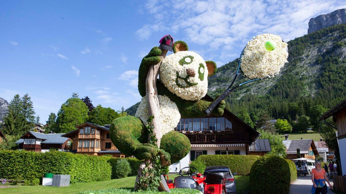 Diese Narzissenfigur zeigt einen Pandabären, der an einem Stamm hinaufklettert und einen Tennisschläger mit Tennisball in der Hand hält. Im Hintergrund sieht man das schöne Ausseerland. | © Narzissenfestverein_Sima